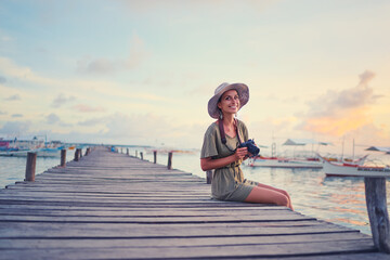 Photography and travel. Young woman in hat holding camera sitting on wooden fishing pier with beautiful tropical sea view.