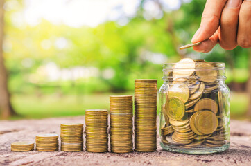 investor business man hand putting coins in jar with money row stack on wood table with blur nature...
