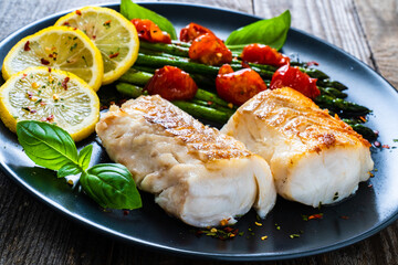 Fish dish - fried cod fillet with asparagus and cherry tomatoes served on black plate on wooden table
