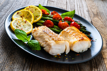Fish dish - fried cod fillet with asparagus and cherry tomatoes served on black plate on wooden table
