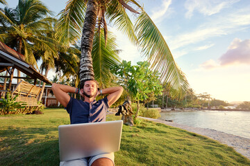 Work and vacation. Young man working on laptop computer on the tropical beach under the palm tree.