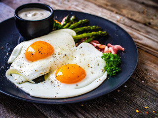 Tasty breakfast. Sunny side up eggs with green asparagus, fried bacon and garlic dip served on black plate on wooden table