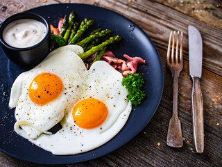 Tasty breakfast. Sunny side up eggs with green asparagus, fried bacon and garlic dip served on black plate on wooden table