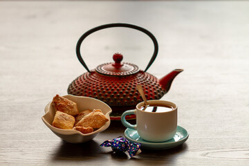 A cup of tea with cookies on the background of a red teapot standing on a dark table.