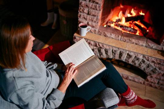 Cozy home. Pretty young woman is reading book near the fireplace.