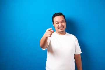 Portrait of young Asian man in white t-shirt smiling and pointing forward, looking at camera. Choosing someone concept with selective focus, against blue background