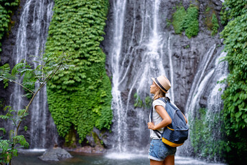 Tropical traveling. Young woman in hat with rucksack enjoying waterfall view.