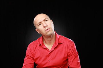 Headshot of bald adult man in red shirt with funny emotions isolated on black background with copy space.