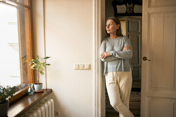 Indoor shot of serious sad middle aged female in casual clothes standing at doorway keeping arms...