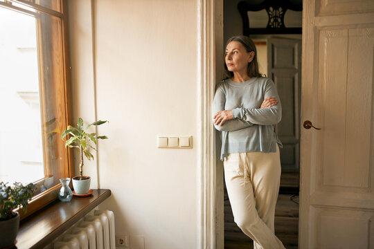 Indoor shot of serious sad middle aged female in casual clothes standing at doorway keeping arms crossed on her chest, looking through window, being upset while staying at home during quarantine