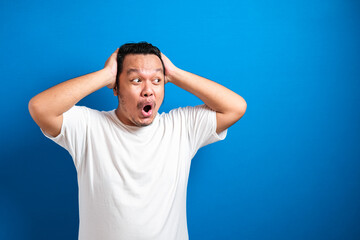 Naklejka premium Fat Asian man in white t-shirt shows funny shocked and surprise expression against blue background