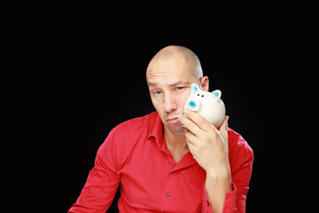 Headshot of adult caucasian bald man in red shirt his holding ceramic piggy bank isolated on black background.