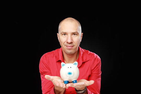 Headshot of adult caucasian bald man in red shirt his holding ceramic piggy bank isolated on black background.