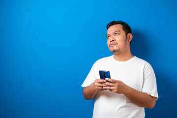 Fat Asian man in white t-shirt, thinking something while chat messaging on his smart phone, half...