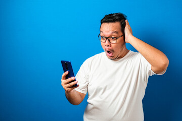 Fat Asian guy wearing a white t-shirt looks surprised at the good news he received from his smartphone. Men show shocked movements with bulging eyes while rolling down his glasses on a smartphone