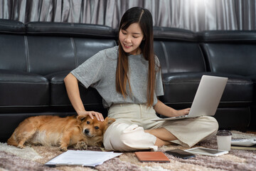 Pretty asian woman working remotely from home using laptop sitting on the couch or sofa in living room for work online with pet puppy cute dog and guardian, work life balance concept.