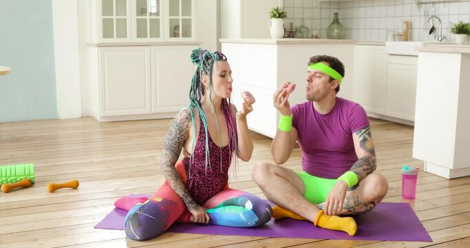 Copule of man and woman in sportswear enjoying of eating donuts sitting on fitness mat at home. Diet, junk food, spot concept. Humor, mem, joke, parody, fun behaviour. Addiction of bakery food.