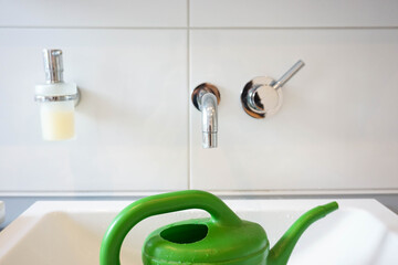 A hand grips a filled watering can in a bathroom sink to water indoor plants.