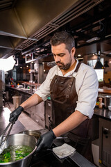 Busy bearded chef in apron standing at stove and boiling broccoli in pot at kitchen