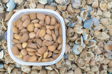 Peeled almonds in a square transparent box. In the background, the shell is in blur. View from above, flat lay. Copy space, place for text.