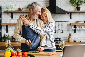 Beautiful mature couple hugging while cooking vegetable salad at kitchen table - obrazy, fototapety, plakaty
