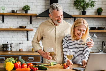 Senior couple watching in laptop and drinking wine while cooking dinner meal