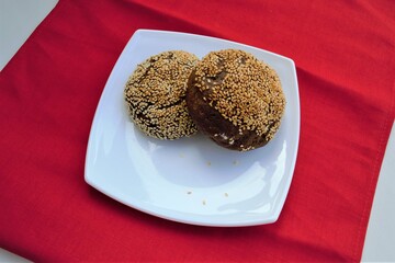 Composition of homemade rye buns with sesame seeds on top (and several seeds nearby) on a square glossy plate standing on bright red linen cloth