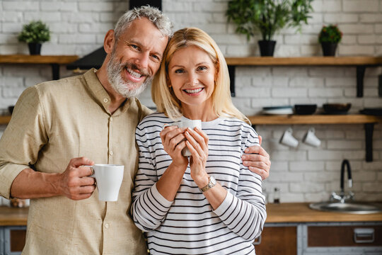 Beautiful Middle Aged Couple Drinking Tea Or Coffee At Home While Looking At Camera