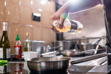 Close-up of unrecognizable chef standing at stove and pouring oil into cooking pan at commercial kitchen