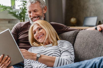 Close up of smiling middle aged couple using digital tablet on couch at home interior