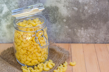 Close-up of Uncooked Macaroni in a glass jar placed on the wooden table with cement wall background , selective focus. - 355907630
