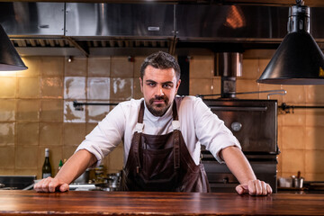 Portrait of confident handsome bearded chef in leather apron standing at wooden counter in open kitchen