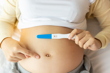 Happy pregnant woman holding pregnancy test pointing finger to beautiful belly that pregnant girl has fetus inside. Young Mother waiting newborn baby prenatal, pregnancy, motherhood, expecting concept