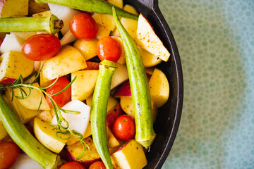 Top view of an iron pan with raw, colorful, healthy and organic food like potato, sweet potato, tomato and okra for a meal with a green background.