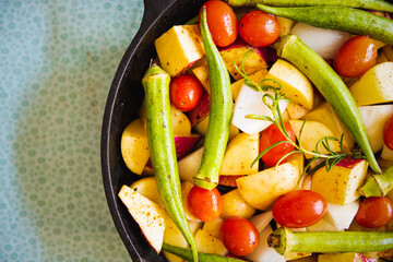 Top view of an iron pan with raw, colorful, healthy and organic food like potato, sweet potato, tomato and okra for a meal with a green background.