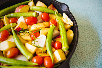 Top view of an iron pan with raw, colorful, healthy and organic food like potato, sweet potato, tomato and okra for a meal with a green background.