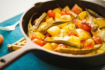 Top view of an iron pan with cooked, colorful, healthy and organic food like potato, sweet potato, tomato and okra for a meal with a green background.