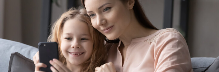 Horizontal panoramic view of loving young Caucasian mother and small daughter have fun browsing smartphone together, happy mom and little girl child watch video use cellphone gadget at home