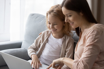 Happy young Caucasian mom and small daughter relax on sofa watch video on laptop together, smiling millennial mother and little daughter have fun browsing internet learnings studying on computer