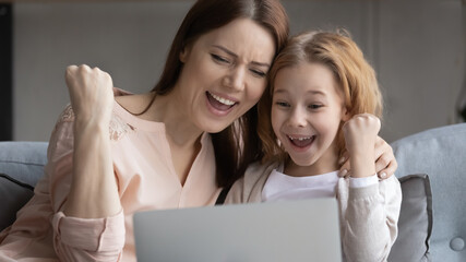 Excited young Caucasian mother and little daughter triumph win online application game on laptop, overjoyed mom or nanny have fun feel euphoric gain victory in online lottery on computer