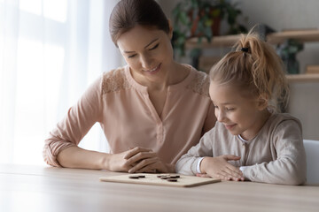 Smiling caring young mother teach little daughter play wooden chess or draughts, happy playful Caucasian mom or nanny engaged in educational game activity at home together, entertainment concept