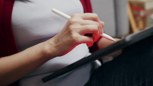 Close up of Asian Female Freelancer or Graphic Designer working on a digital tablet using stylus pen drawing on a graphic tablet sitting at her workplace in the living room at home office.