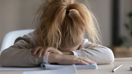 Tired little schoolgirl sit at desk fall asleep feel lazy unmotivated to study, exhausted small...