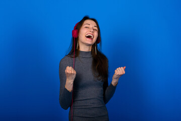 Happy satisfied girl istanding listening favorite music with headphones, closed eyes and smiling, Indoor studio shot, isolated on blue background