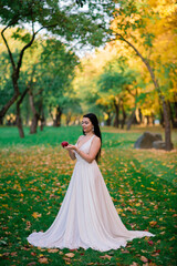 Obraz na płótnie Canvas Young beautiful brunette female in a long white dress in autumn park.