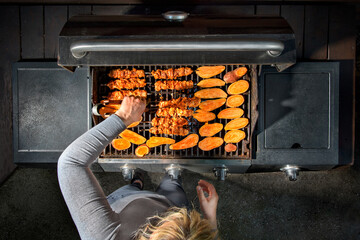 Woman using a gas grill in an outdoor kitchen. Shish kebab on skewers and sweet potato.
