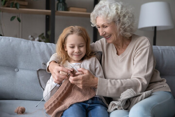 Caring mature grandmother sit rest on sofa have fun teach little granddaughter knitting with...