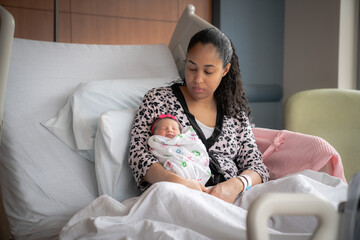 An African American mixed race mother in a pink animal print bathrobe sits on a hospital bed...