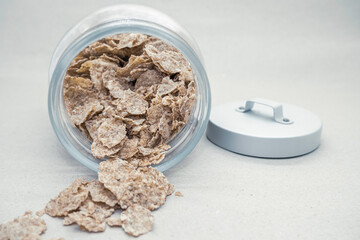 Whole grain flakes falling out of a glass jar next to a metal cap on a gray background close-up