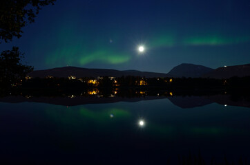 majestic aurora borealis, northern light over calm mirror lake at night with buildings in the background